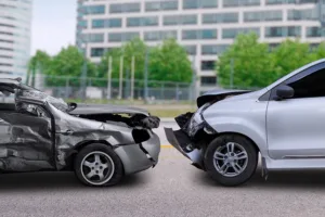 Two cars with significant front-end damage face each other on a road, appearing to have been involved in a head-on collision—an image often seen in car accidents. A blurred building and trees are visible in the background.