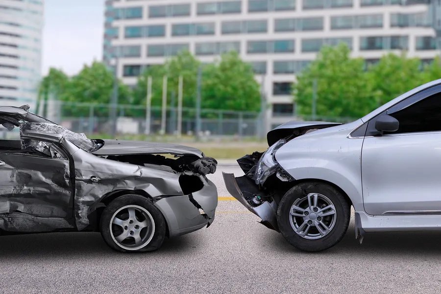 Two cars with significant front-end damage face each other on a road, appearing to have been involved in a head-on collision—an image often seen in car accidents. A blurred building and trees are visible in the background.