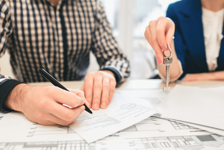 A person signs a document while another holds a set of keys, suggesting a real estate or property transaction. Lawyers may oversee such agreements, with papers and architectural drawings visible on the desk.