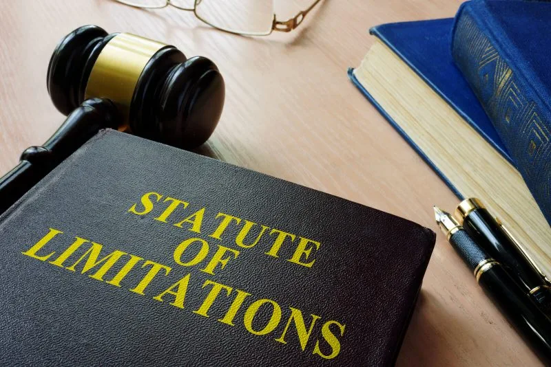 A book titled "Statute of Limitations" sits on a desk next to a judge's gavel, eyeglasses, a pen, and another closed book—a fitting scene for lawyers or anyone dealing with car accidents seeking legal guidance.