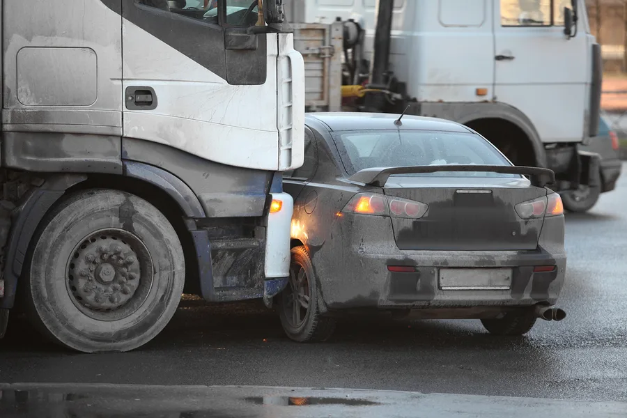 A large truck and a black car with dirty exteriors are involved in a collision at an intersection, with the truck’s front bumper pressed against the side of the car—an incident that may require a personal injury attorney.