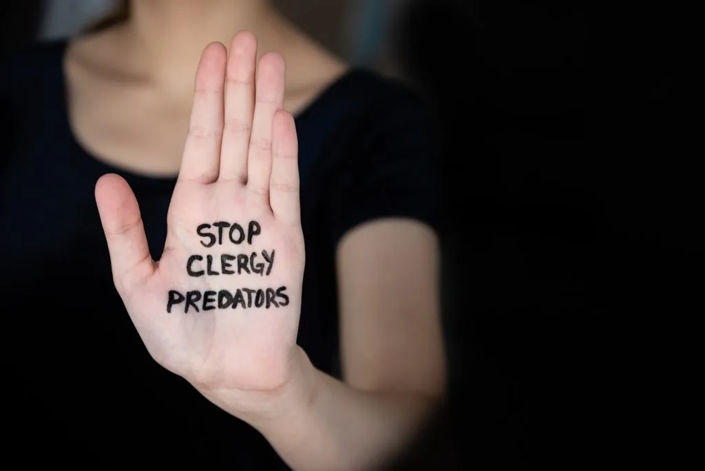 A person holds up their hand with "STOP CLERGY PREDATORS" written in black marker on their palm, conveying a message against clergy abuse. The dark background and hidden face evoke the anonymity faced by many personal injury survivors seeking justice.