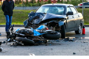 A heavily damaged black car and a wrecked blue motorcycle lie on a road after a collision, with debris scattered around. A person stands nearby, and a fire extinguisher is on the ground.