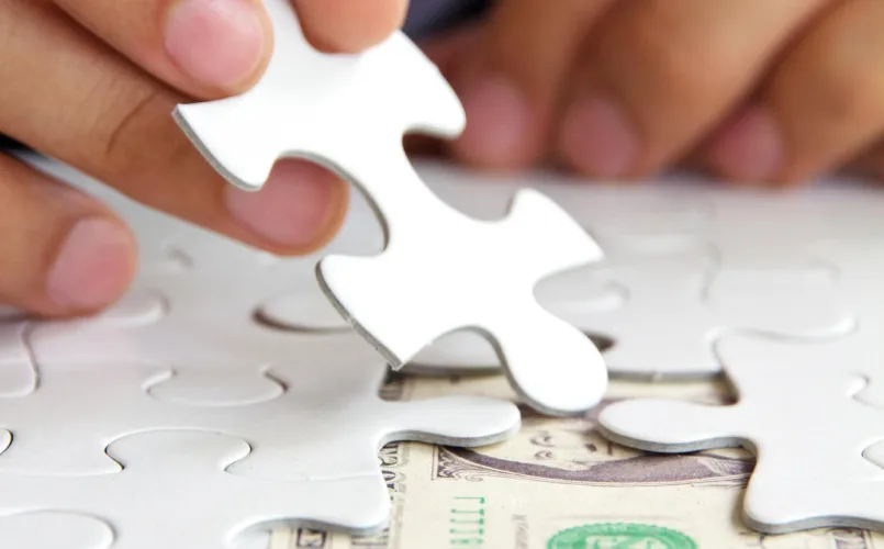 Close-up of hands placing a white puzzle piece into a nearly completed puzzle, partially revealing a U.S. dollar bill underneath—symbolizing how personal injury attorneys help clients fit the pieces together after an accident.