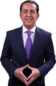A personal injury attorney in a navy blue suit, white shirt, and purple tie stands smiling with his hands clasped in front of him against a plain, light background.