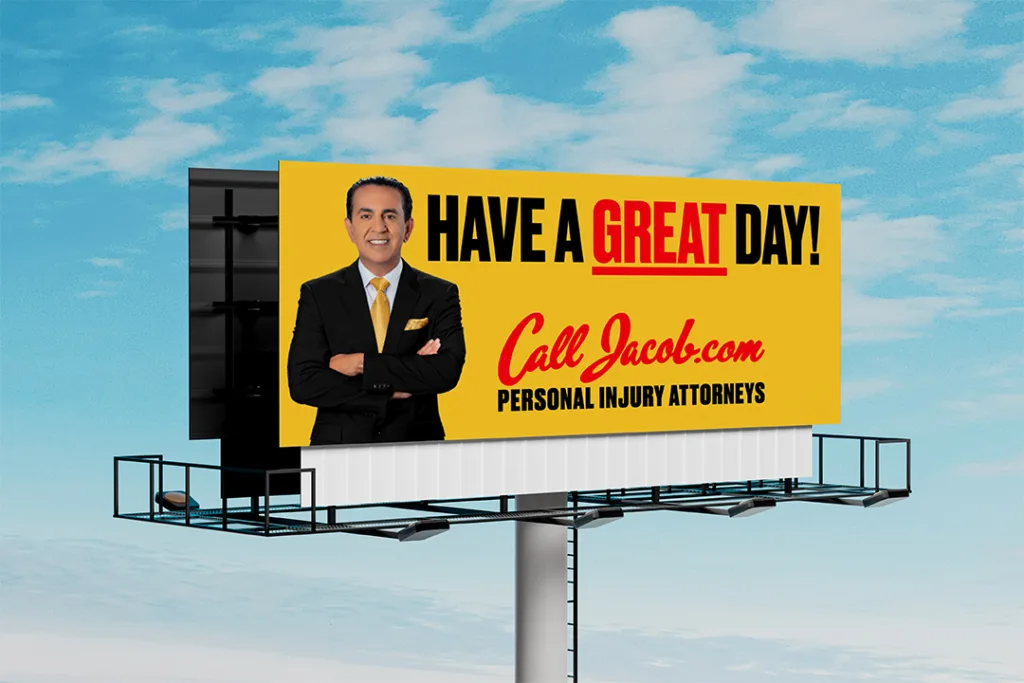 A billboard features a man in a suit with folded arms, and text that says, "HAVE A GREAT DAY! Call Jacob.com PERSONAL INJURY ATTORNEYS" against a yellow background, promoting help for car accidents. The sky is blue with some clouds.