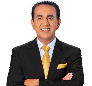 A man in a black suit, white shirt, and yellow patterned tie stands with arms crossed, smiling at the camera against a light background. He has dark hair slicked back and is wearing a matching yellow pocket square.