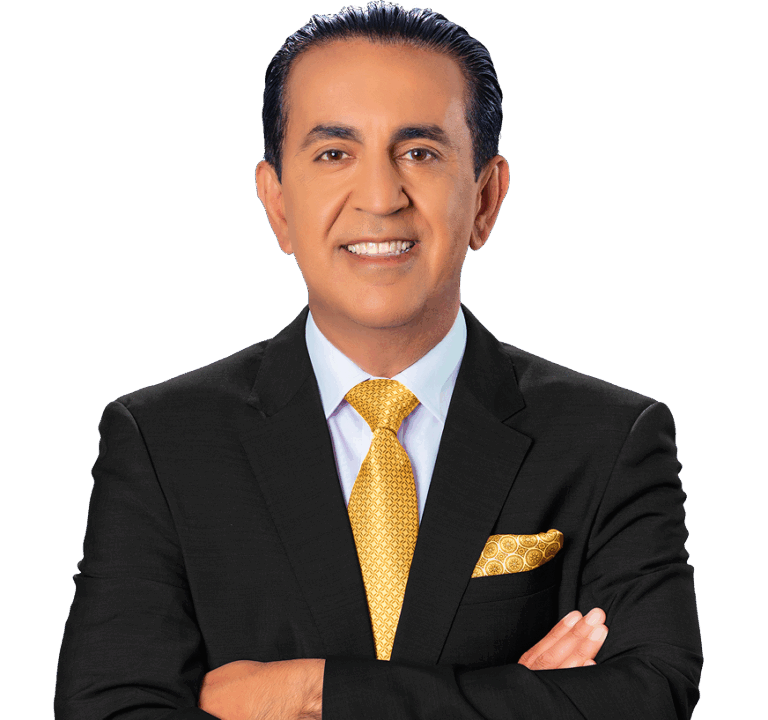 A man in a black suit, white shirt, and yellow patterned tie stands with arms crossed, smiling at the camera against a light background. He has dark hair slicked back and is wearing a matching yellow pocket square.