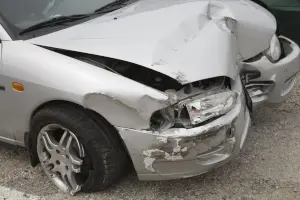 A silver car with significant damage to its front bumper and hood, indicating a recent collision. The front right headlight is broken, and another vehicle is visible next to it.