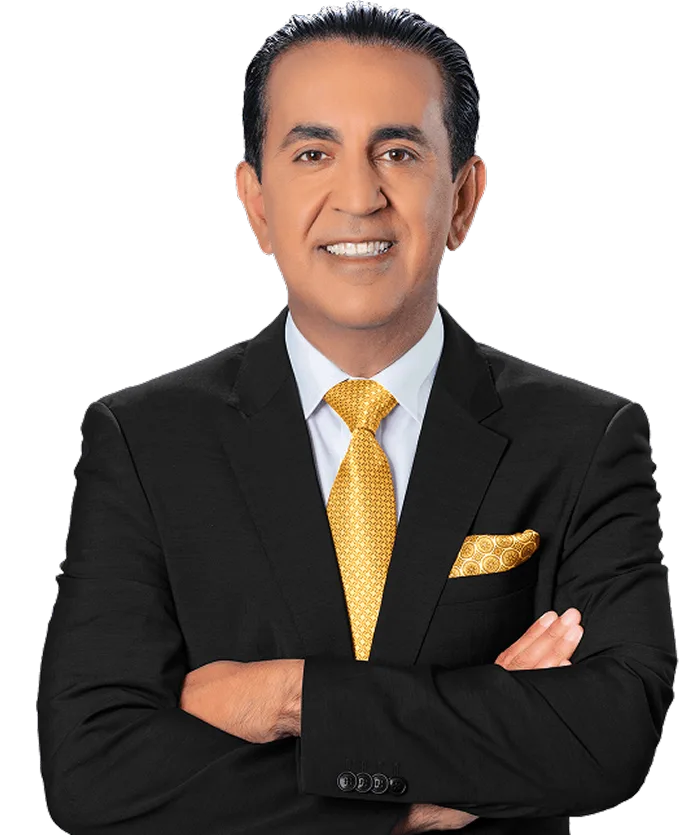 A man in a black suit with a white shirt and a yellow patterned tie stands with arms crossed, smiling at the camera. He has dark hair neatly styled and a matching yellow pocket square. The background is plain and light-colored.