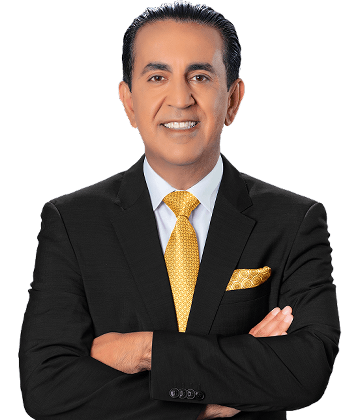 A man in a black suit with a white shirt and a yellow patterned tie and pocket square stands with his arms crossed, smiling at the camera against a plain light background.