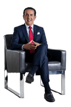 A man in a dark suit, red tie, and black shoes sits in a modern armchair with his hands clasped, facing forward and smiling.