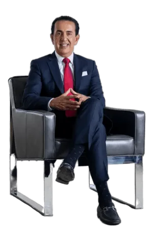 A man in a dark suit, red tie, and black dress shoes sits in a modern black armchair, hands clasped, smiling at the camera against a plain white background.