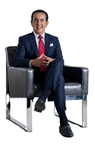 A man in a dark suit, red tie, and black dress shoes sits in a modern black armchair, hands clasped, smiling at the camera against a plain white background.