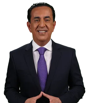 A man in a dark suit, white shirt, and purple tie stands against a plain background, smiling confidently with his hands together—truly looking like el mero mero.