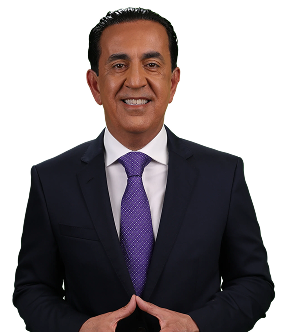 A man in a dark suit, white shirt, and purple tie stands against a plain background, smiling confidently with his hands together—truly looking like el mero mero.