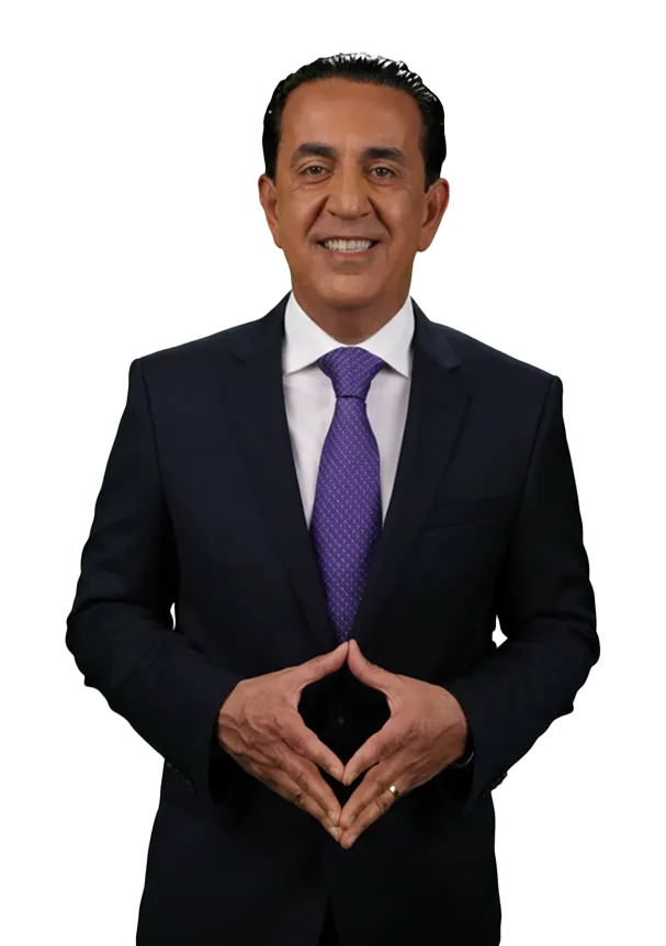 A man in a dark suit, white shirt, and purple tie stands smiling with his hands forming a triangle shape in front of him, exuding confidence like el mero mero. The background is plain white.