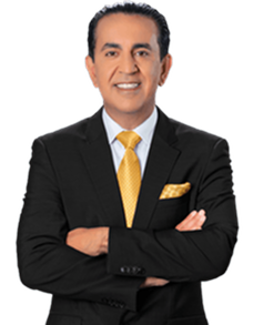 A man in a black suit, white shirt, and yellow tie stands with arms crossed, smiling at the camera. He has short dark hair and a pocket square matching his tie. The background is plain and light-colored.