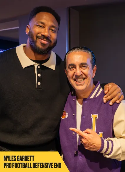 Two men pose and smile for a photo indoors. One wearing a black and tan collared shirt, the other in a purple and white varsity jacket points at his companion. Text reads: "MYLES GARRETT PRO FOOTBALL DEFENSIVE END".