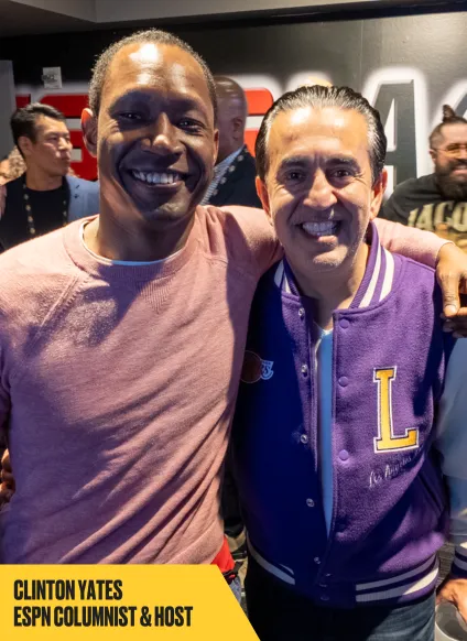 Two men smiling and posing together at an indoor event. The man on the left wears a pink sweater, and the man on the right wears a purple "L" varsity jacket. Text at bottom reads: "CLINTON YATES ESPN COLUMNIST & HOST.