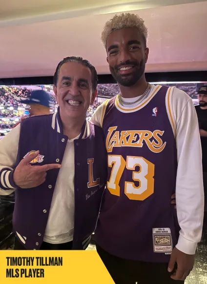 Two men are smiling and posing together indoors. One is wearing a purple Lakers jacket and pointing at the other, who is wearing a Lakers jersey with the number 73. Text reads, "Timothy Tillman MLS player.