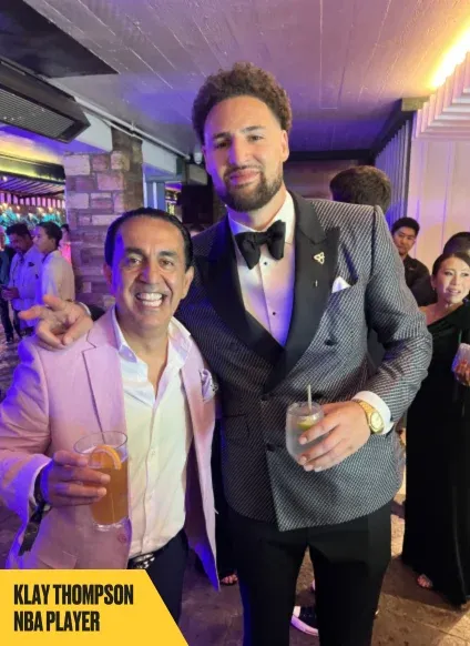 Two men pose together at an indoor event, both smiling and holding drinks. The taller man is in a black and white tuxedo, and the other is in a light-colored suit. A yellow label reads "Klay Thompson NBA Player.