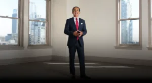 A man in a dark suit with a red tie stands confidently in a large, empty room with tall windows showing a cityscape of modern skyscrapers in the background.