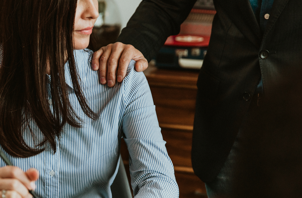 A person in a suit places their hand on the shoulder of a woman in a blue striped shirt, who looks away.