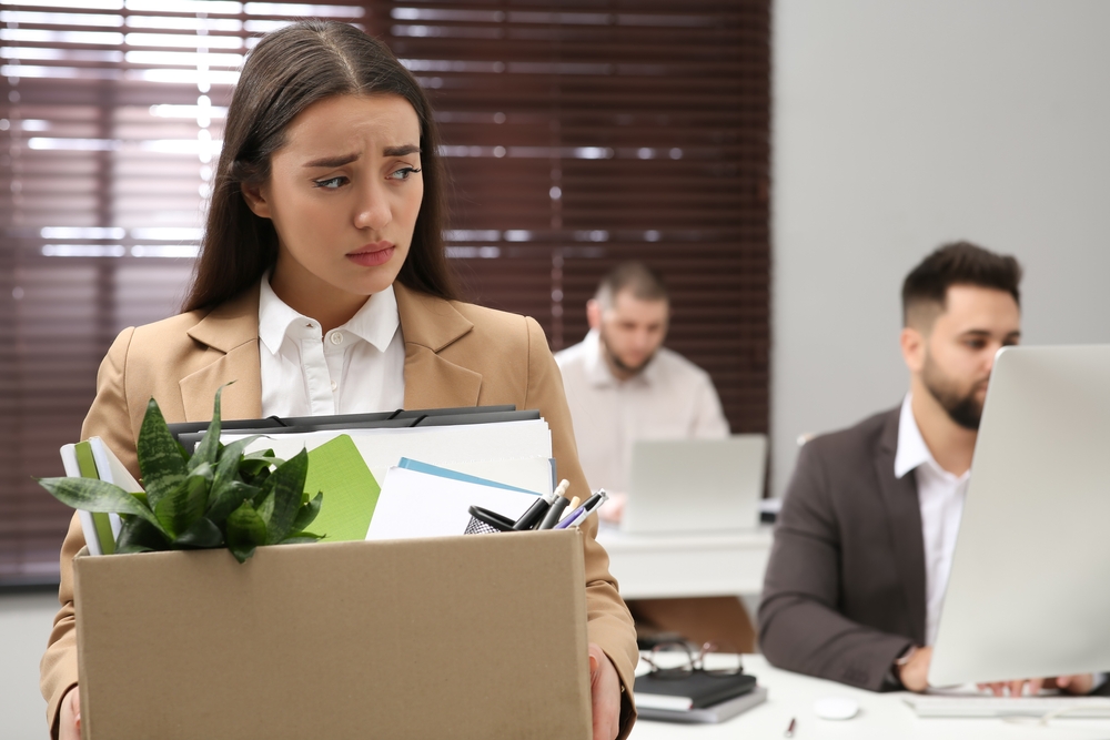 A sad woman holding a box of office supplies, likely just laid off, stands in an office while two colleagues work at computers in the background.
