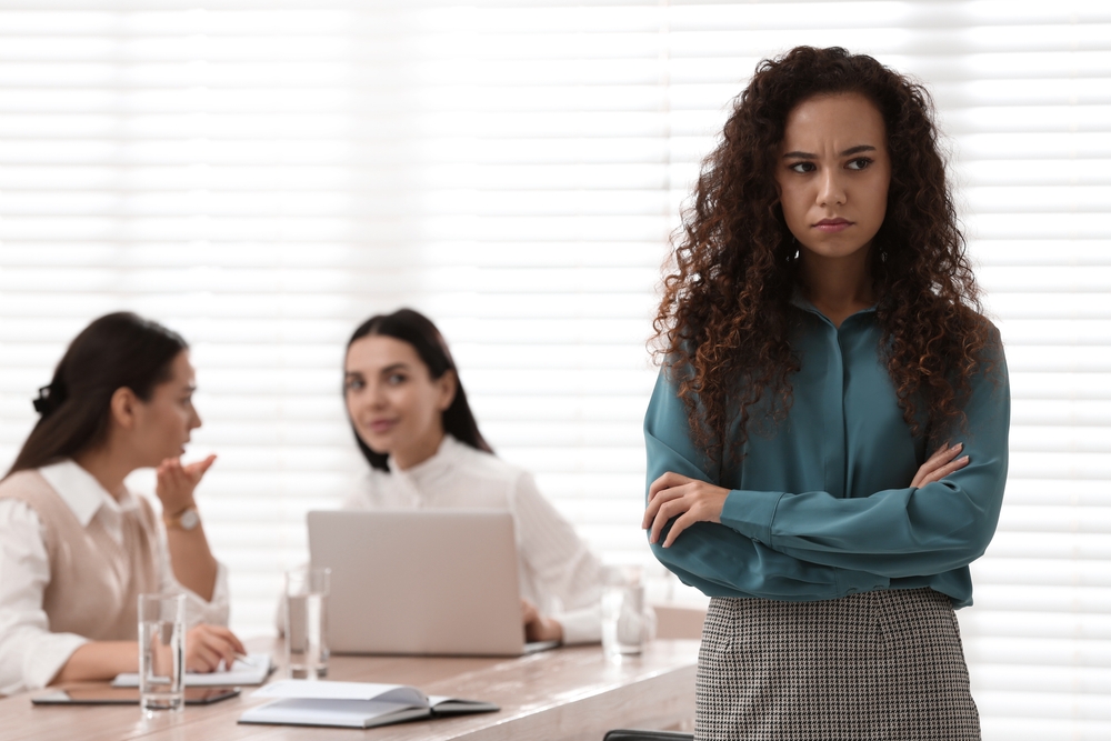 A woman stands with arms crossed, looking upset, while two colleagues sit at a table behind her, whispering and glancing in her direction.