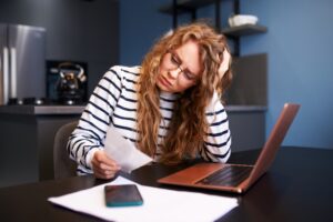 A woman with long curly hair and glasses, wearing a striped shirt, sits at a table with a laptop, smartphone, and papers, looking concerned as she reads a receipt in a modern kitchen.